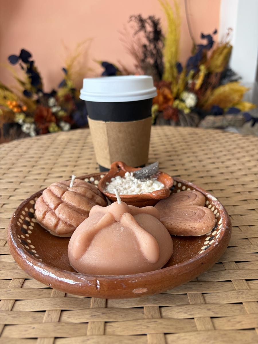 Velas con forma de concha, pan de muerto y oreja en un plato de barro, junto a un café